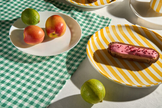 Sunlit table with pink eclair, fresh fruit on green gingham tablecloth