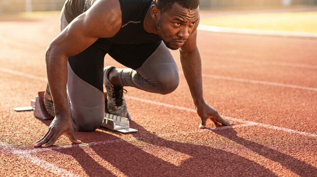 Muscular professional sprinter focuses intensely while setting up on starting blocks on a red track during sunrise. High-quality athletic training and preparation stock.