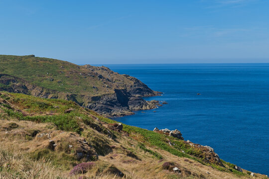 A vibrant coastal scene with rolling green and brown hills meeting a deep blue ocean under a clear sky. The sunny day highlights the rugged cliffs - Botallack Mine & Engine House, Cornwall, UK