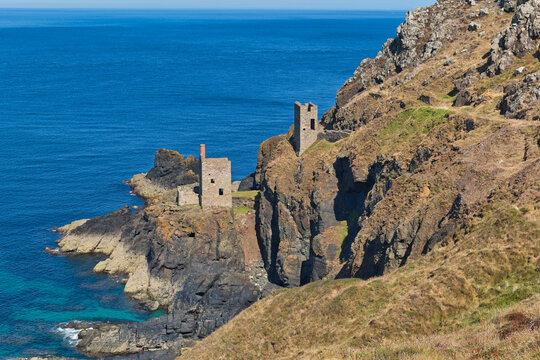 Historic stone engine houses cling to a rugged cliff overlooking a vibrant blue sea. This dramatic coastal scene is bathed in sunlight - Botallack Mine & Engine House, Cornwall, UK