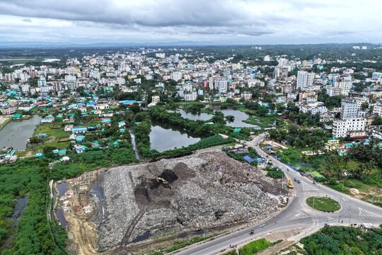 Aerial view of a large landfill site with an excavator working on waste, surrounded by lush vegetation, ponds, and the urban cityscape of Cox's Bazar, Chittagong Division, Bangladesh.