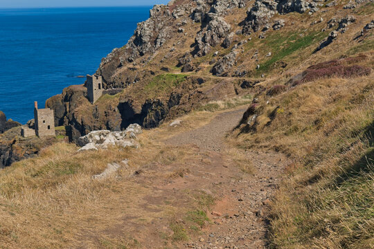 A scenic coastal path stretches along rugged cliffs with historic stone engine houses overlooking the vibrant blue sea - Botallack Mine & Engine House, Cornwall, UK