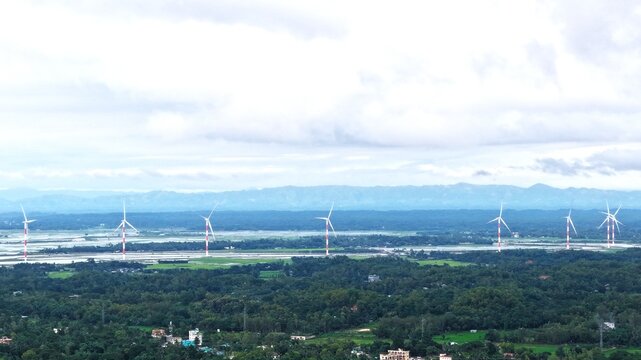 Aerial view of the wind power plant with white and red turbines standing in a lush green landscape under a cloudy sky in Cox's Bazar, Chittagong Division, Bangladesh.