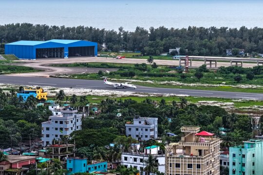 Aerial view of Cox's Bazar International Airport featuring a plane on the runway, a large blue hangar, and surrounding urban buildings in Cox's Bazar, Chittagong Division, Bangladesh.