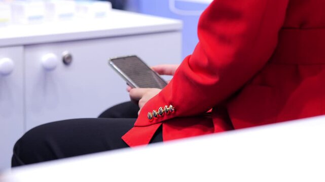 Person in red jacket holds smartphone. White cabinet with knobs behind. Focus on hands and device. Modern interior setting. Minimalist composition with soft lighting