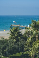 Tureckie wybrzeże Morza Śródziemnego – plaża, palmy i turkusowe morze. Widok na słoneczne wybrzeże Turcji nad Morzem Śródziemnym. Turkusowa woda, kamienista plaża, palmy, pomost hotelowy. © MarcinRoj.Fotografia