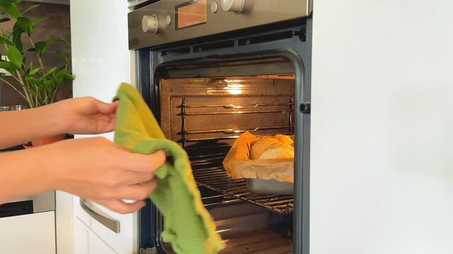 Hands in green oven mitts take tray of freshly baked bread rolls out of oven. Home baking and domestic culinary process concept.