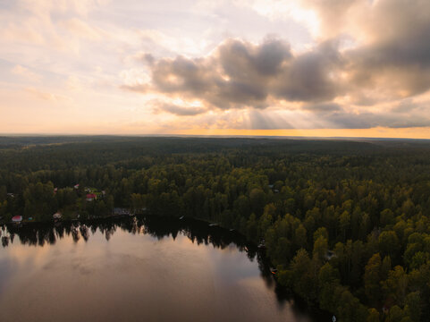 Aerial view of Siikajarvi lake surrounded by dense evergreen forests during a golden sunset with sun rays breaking through the clouds in Siikajarvi, Uusimaa, Finland.