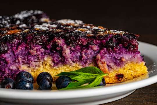 Slice of homemade blueberry cake with fresh berries and mint. Closeup of delicious slice of juicy berry pie or cake decorated with powdered sugar, fresh blueberries, and green mint on white plate
