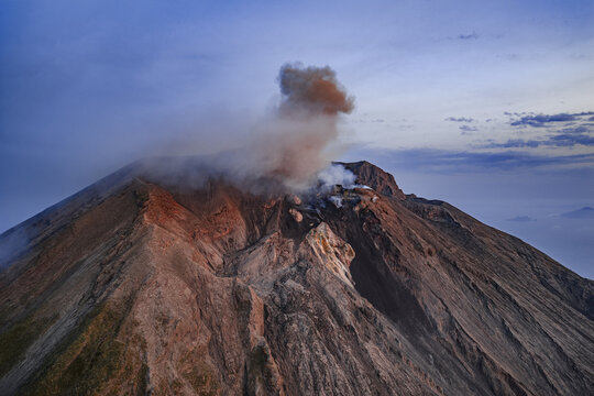Aerial view of Stromboli volcano crater with a plume of smoke and ash rising into the twilight sky over the rugged volcanic landscape in Lipari, Sicily, Italy.