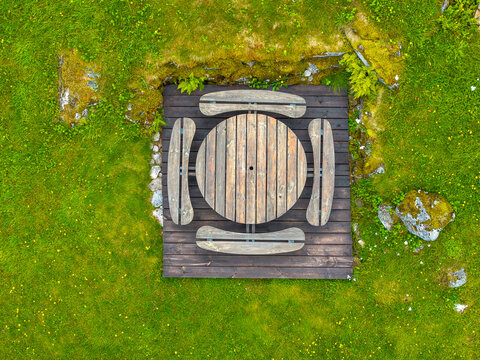Aerial view of a circular wooden picnic table set on a dark timber deck surrounded by lush green grass and mossy stones in Sylte, More og Romsdal, Norway.