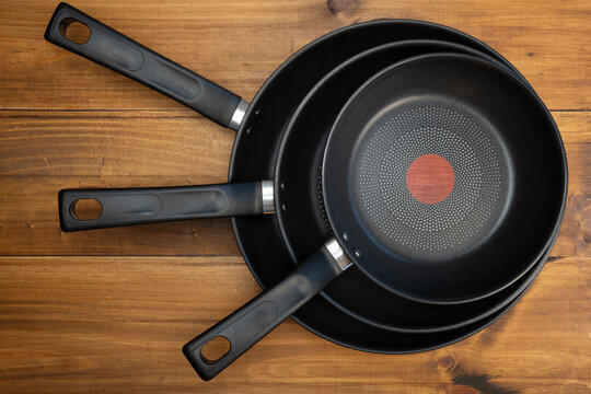 Three black non-stick frying pans stacked on a rustic wooden kitchen table, top view showing different sizes and handles