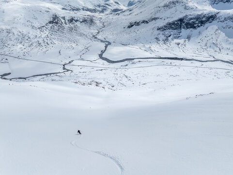 Aerial view of a lone skier carving a path down a vast, pristine snow-covered mountain slope above a winding river valley in Valldal, More og Romsdal, Norway.