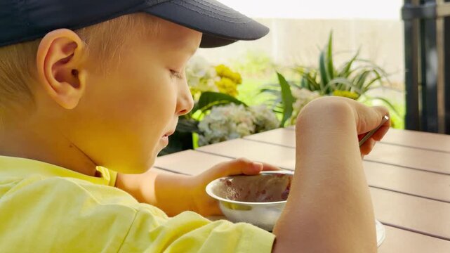 A joyful boy eats food with a spoon outdoors at a wooden table. He feels happy and content enjoying a cozy summer meal.