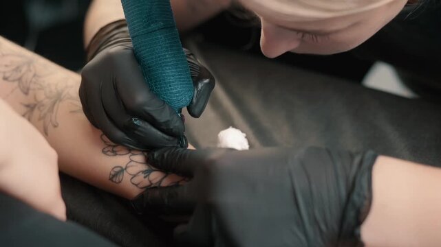 Professional tattoo artist creating a tattoo on a woman's forearm using a tattoo machine in a modern studio. Client relaxing during the tattooing procedure.