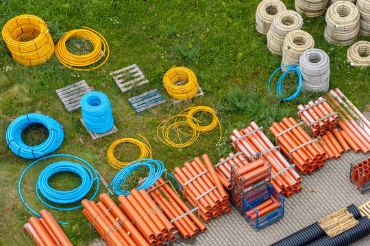 Aerial view of various plastic construction pipes, including yellow, blue, and orange coils and straight sections, stored on a grassy field and paved area in Poland.