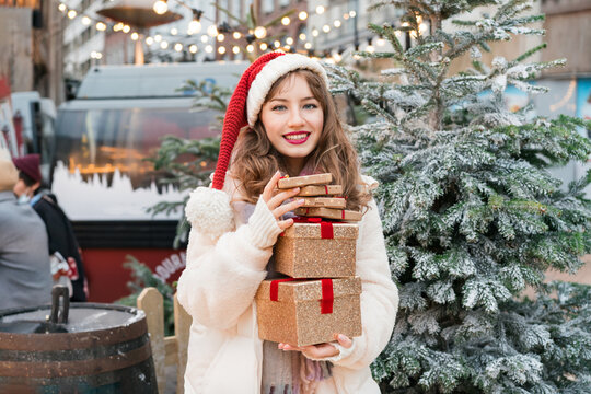 Young woman in Santa hat with with a tower of gift boxes on traditional city street Christmas market. Happy woman choosing Christmas gifts. Merry Xmas Happy New Year festive shopping, sales, discount.
