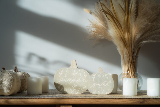 Cozy warm autumn home interior - white knitted pumpkins, wooden statuette, candles, dried flowers on the wooden console with white wall background. Scandinavian minimalist hygge home fall decor.