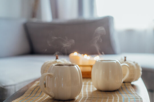 Autumn fall mood composition for hygge home decor. Hot tea or coffee drink in pumpkin's shaped cups, pumpkin decor and burning candles on the coffee table. Festive autumn, fall mood home decor