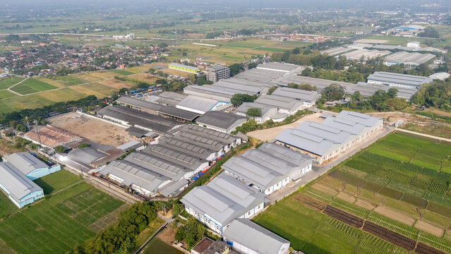 Aerial view of an industrial complex with large metal-roofed warehouses surrounded by lush green rice fields and rural landscape in Central Java, Indonesia.