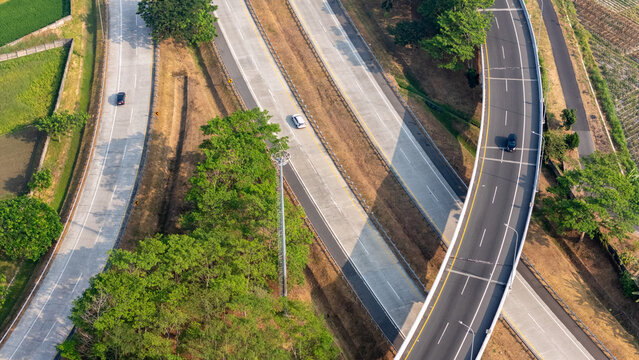 Aerial view of a highway interchange with cars driving on concrete lanes, surrounded by lush green trees and agricultural fields in Central Java, Indonesia.