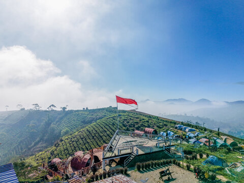 Aerial view of a viewing platform with the Indonesian flag set amidst lush green tea plantations and camping tents on a misty mountain slope in Indonesia.