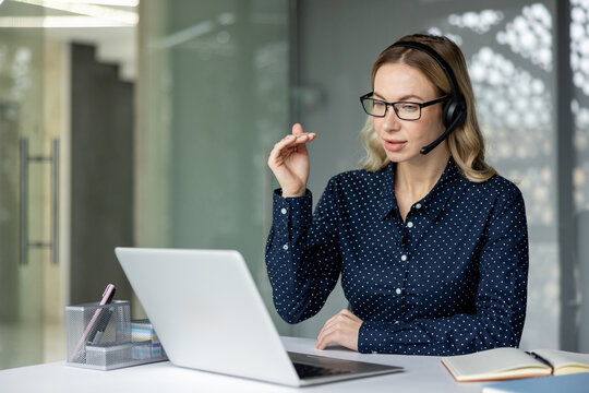 Woman wearing headset and glasses, communicating on a video call using a laptop, working remotely from an office desk while engaged in customer support or online consultation