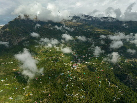 Aerial view of Spiti valley with a village nestled among green mountain slopes, dense forests, and scattered clouds in Lahaul and Spiti, Himachal Pradesh, India.