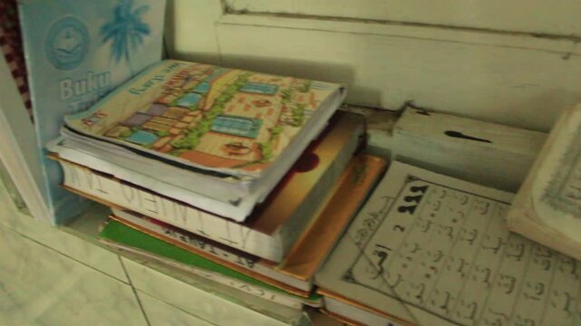 Handheld view of a messy stack of books and Qurans beside a window inside a building