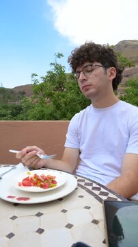 Marrakech, morocco. August 9, 2026. Caucasian man with curly hair and glasses eating a fresh tomato salad outdoors at a typical restaurant in marrakech, morocco