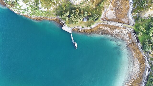 Aerial view of Aroya island featuring a wooden pier extending into the turquoise waters of the fjord, surrounded by rocky cliffs and lush green vegetation, Alta, Finnmark, Norway.