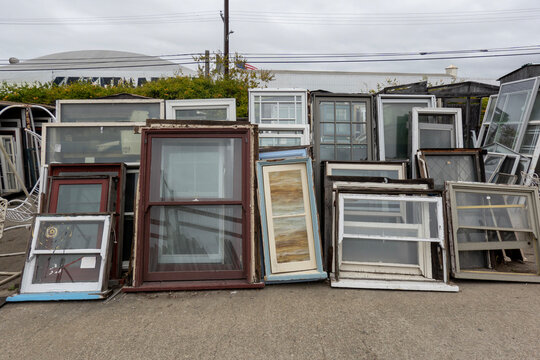 used windows-An assortment of various salvaged  vintage and used window frames in different sizes and wood finishes are stacked vertically outside.