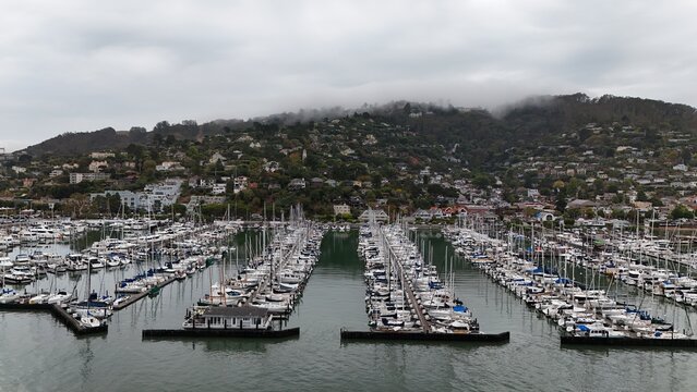Aerial view of a marina filled with numerous sailboats docked in rows, with the hillside town of Sausalito and fog-covered hills in Sausalito, California, United States.