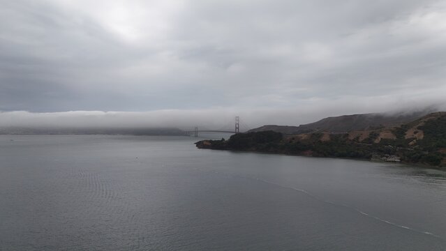 Aerial view of Golden Gate Bridge partially obscured by low-hanging fog over the bay with surrounding coastal hills under a cloudy sky in Sausalito, California, United States.