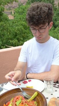 Marrakech, morocco. August 9, 2026. Young tourist with glasses eating a traditional moroccan vegetable tagine. A culinary experience on a terrace with a lush green backdrop