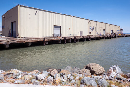 transit shed or a freight shed-a long transit shed building sits alongside a calm body of water with a rocky shoreline in the foreground.
