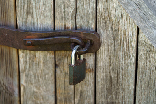 Old rusty padlock hanging on a forged metal latch of a wooden door. Close-up shot capturing vintage rural life and antique rustic details.