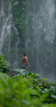 Handsome man with raised arms under powerful waterfall, feeling energy in tropical jungle nature