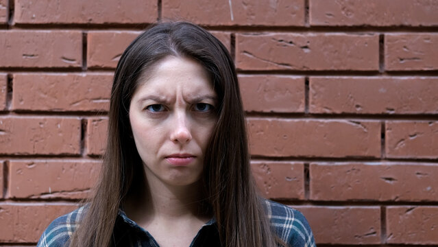 Serious young woman with long brown hair standing against weathered red brick wall, staring directly at camera with furrowed brows and neutral posture in outdoor urban portrait