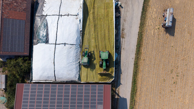 Aerial view of a livestock farm featuring buildings with solar panels, a green tractor working on a large silage pile, and adjacent agricultural fields in Mediglia, Lombardy, Italy.