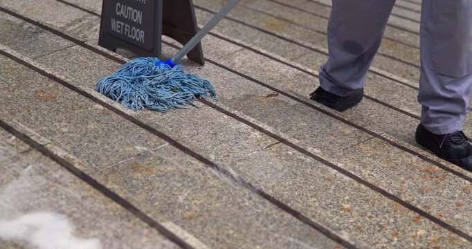 Professional cleaner mopping granite stairs with blue mop, caution wet floor sign in background, facility maintenance and hygiene service in public area, janitorial work.