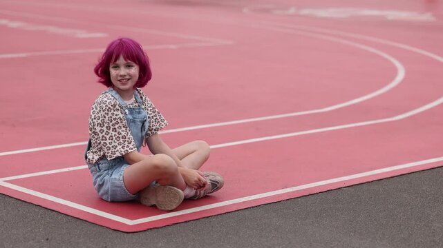 Young child with short pink hair and beige hat sits on red outdoor running track near white curved lane lines, wearing denim overalls with sunglasses clipped to pocket and leopard print shirt