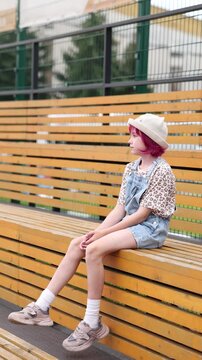 Child with pink hair sits alone on outdoor wooden bleachers wearing light hat, patterned short sleeve shirt, and denim overalls, relaxed neutral expression, casual sneakers and white socks visible