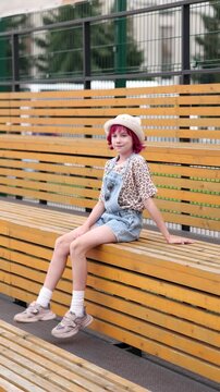 Child with pink hair sits alone on outdoor wooden bleachers wearing light hat, patterned short sleeve shirt, and denim overalls, relaxed neutral expression, casual sneakers and white socks visible
