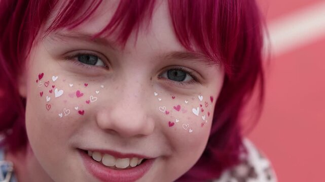 Closeup portrait of smiling child showing upper face and mouth, bright pink dyed hair with bangs framing forehead, tiny heart stickers in white and pink scattered across both cheeks, freckled skin