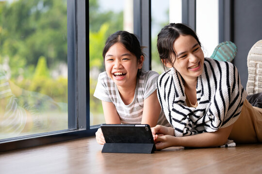 Laughing woman and girl using tablet beside a window at home, sharing happiness, technology, togetherness and leisure while relaxing on the floor with cheerful expressions