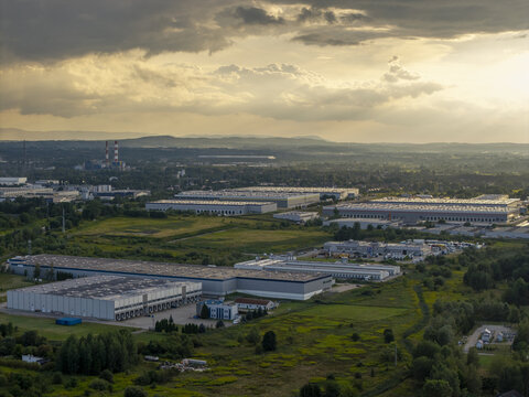 Aerial view of large industrial warehouses and logistics centers surrounded by green fields and trees under a dramatic cloudy sky at sunset in Krakow, Lesser Poland Voivode, Poland.