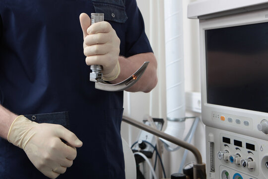 Medical professional in scrubs holds laryngoscope. Anesthesia machine with monitor nearby. Clinical preparation for intubation