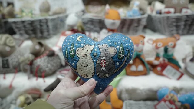 Hand holds heart-shaped blue fabric ornament decorated with two stylized mice, small trees, moon, and polka dot accents, presented forward in foreground while fingers grip bottom edge, ornament