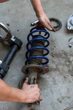 Hands repairing car suspension spring and shock absorber on floor, with coil spring, strut body, worn parts laid out nearby. Dirty fingers grip metal